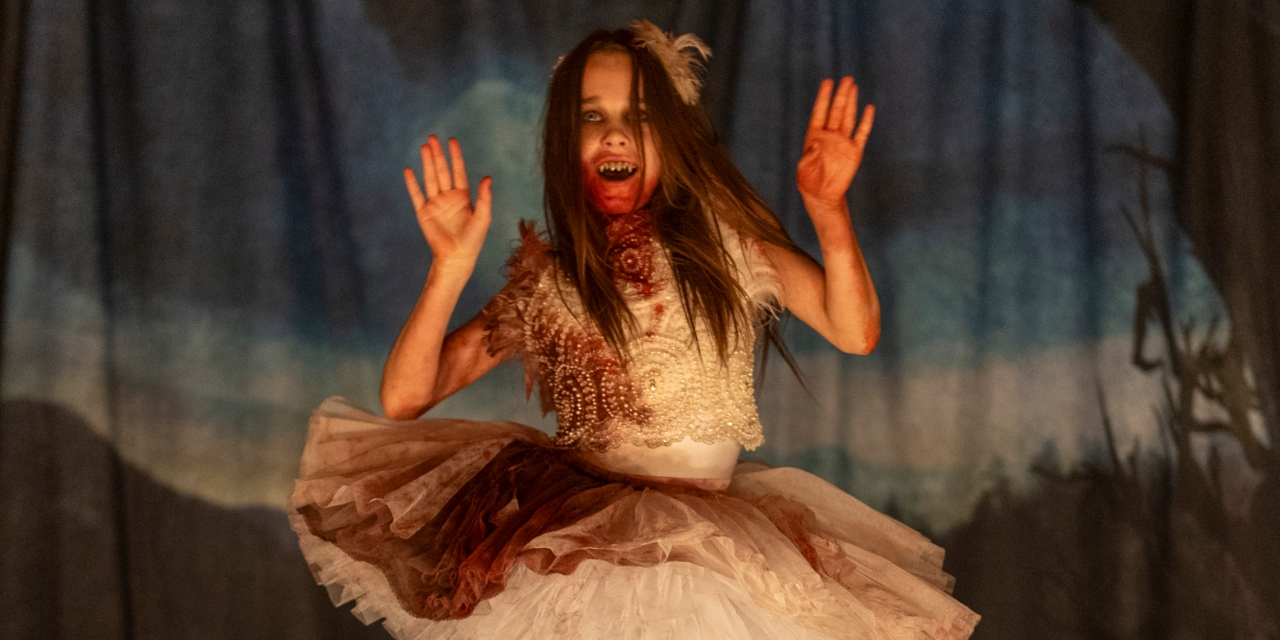 A young girl in a bloodstained white dress with a feathered headdress stands on a stage, holding her hands up and revealing a sinister smile with sharp teeth.